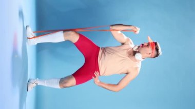 Man, fitness trainer in sportive clothes, bike shorts and cap posing, showing exercise technik with sport expanders on blue studio background. Concept of sportive lifestyle, emotions, retro style. Ad