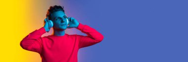 Young handsome man in pink sweater listening to music in headphones over gradient purple yellow background in neon light. Concept of emotions, facial expression, sales, ad, fashion