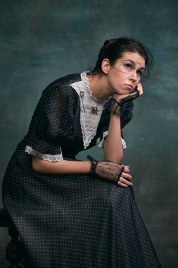 Thoughtful, dreamy look. Beautiful woman in image of Anna Karenina in retro black dress over vintage dark green background. Concept of literature character, history, retro style, comparison of eras