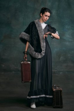 Travelling. Time. Beautiful woman in image of Anna Karenina with retro suitcases over vintage dark green background. Concept of literature character, history, retro style, comparison of eras