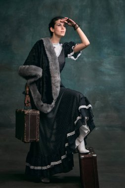 Looking for train. Beautiful woman in image of Anna Karenina with vintage suitcase over vintage dark green background. Concept of literature character, history, retro style, comparison of eras