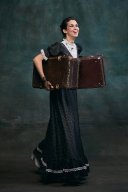 Smiling, holding retro suitcases. Beautiful woman in image of Anna Karenina over vintage dark green background. Concept of literature character, history, retro style, comparison of eras