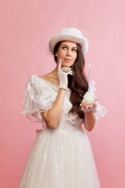 Making wishes. Portrait of beautiful lady in white vintage dress posing with birthday cupcake over pink background. Concept of 19th century, fashion, comparison of eras, history, retro style