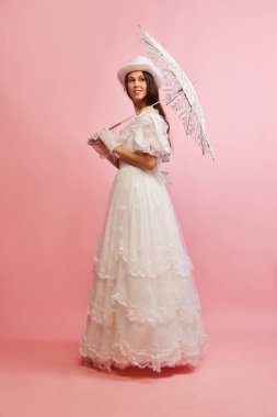 Tender femininity. Portrait of beautiful lady in white vintage dress posing with umbrella over pink background. Concept of 19th century, fashion, comparison of eras, history, retro style