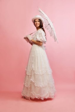 Tender femininity. Portrait of beautiful lady in white vintage dress posing with umbrella over pink background. Concept of 19th century, fashion, comparison of eras, history, retro style clipart