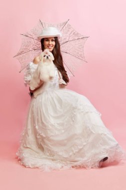 Lovely walk. Portrait of beautiful lady, woman in white vintage dress posing with cute dog over pink background. Concept of 19th century, fashion, comparison of eras, history, retro style