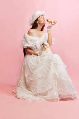 Modern fast food. Portrait of beautiful lady in white vintage dress posing with dog, eating hot-dog over pink background. Concept of 19th century, fashion, comparison of eras, history, retro style