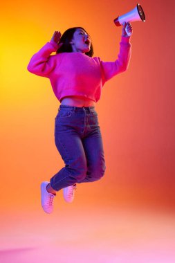 Important news. Young girl, student in pink sweater talking on megaphone in a jump over gradient orange background in neon light. Concept of emotions, facial expression, youth, inspiration, sales, ad