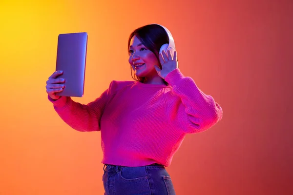 Greetings. Young girl in headphones having online video call on tablet over gradient orange background in neon light. Concept of emotions, facial expression, youth, inspiration, sales, ad