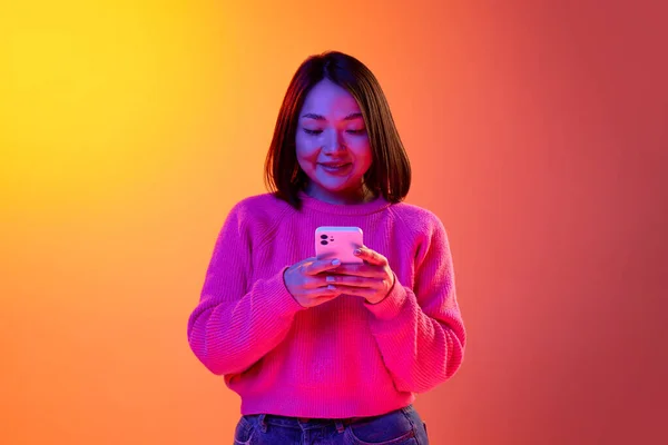 Conversation. Young girl in pink sweater typing text message on mobile phone over gradient orange background in neon light. Concept of emotions, facial expression, youth, inspiration, sales, ad