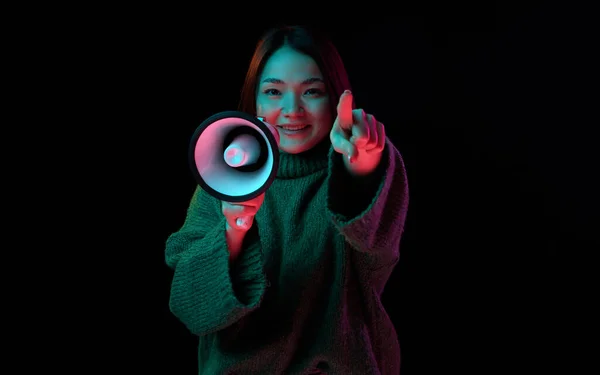 Good, positive news. Young girl in cozy sweater with megaphone pointing at camera with smile over dark background in neon light. Concept of emotions, facial expression, youth, inspiration, sales, ad