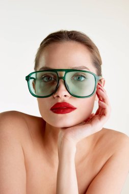 Femininity and sensuality. Young beautiful girl with red lips posing in green sunglases over grey studio background. Concept of natural beauty, skin care, cosmetology, cosmetics, health, fashion