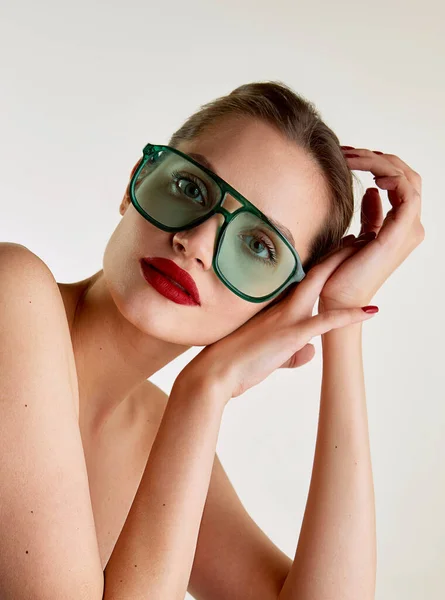 Extravagant style and femininity. Young beautiful girl with red lips posing in green sunglases on grey studio background. Concept of natural beauty, skin care, cosmetology, cosmetics, health, fashion