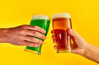 Cheers. Hands clinking green and lager foamy beer glasses over yellow background. Concept of st patricks day celebration, brewery, traditions, alcohol drinks, taste, Irish holiday