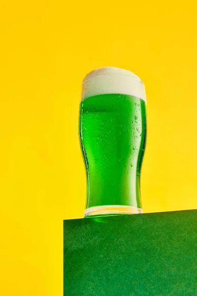 Festival drink. Glass of foamy frothy green beer over bright yellow background. Traditional taste. Concept of st patricks day celebration, brewery, traditions, alcohol drinks, taste, Irish holiday