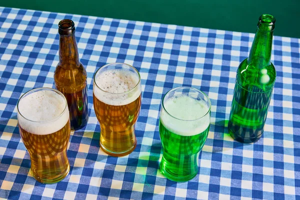 Top view of bottles and glasses with lager and green foamy beer on checkered tablecloth over green background. Concept of st patricks day celebration, traditions, alcohol drinks, taste, Irish holiday