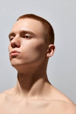 Kısa saçlı, açık tenli, açık gri stüdyo arka planında ciddi bir yüzle poz veren yakışıklı bir gencin portresi. Erkeklerin sağlık ve güzellik anlayışı, vücut ve cilt bakımı, fitness. Vücut sanatı