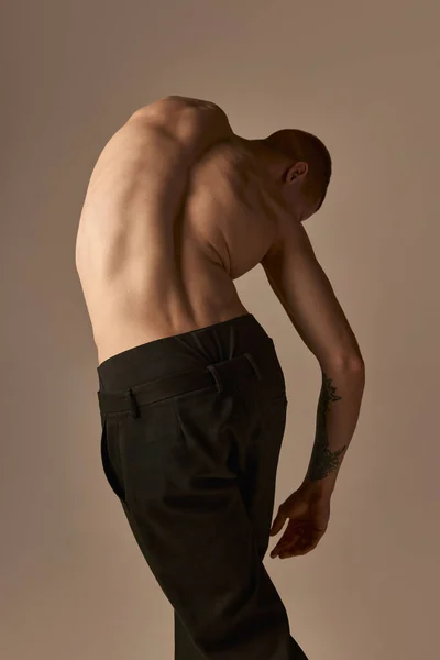 Relief, strong, muscular male back. Young man posing shirtless in pants on light brown studio background. Healthy back, spine. Concept of mens health and beauty, body and skin care, fitness. Body art