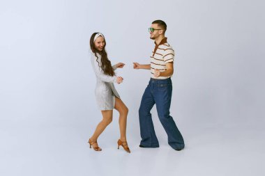 Keyif. Gümüş elbiseli güzel bir kız ve gri stüdyo arka planında izole bir şekilde dans eden şık bir adam. 70 'ler moda, hobi, yaratıcılık, hippi yaşam tarzı, Amerikan kültürü