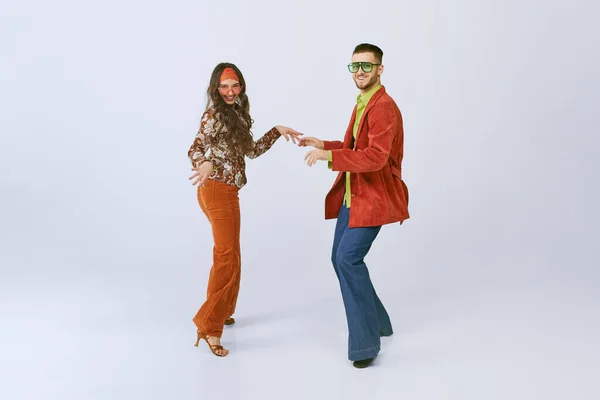 Old school party. Stylish young man and woman in colorful retro outfits dancing disco dance isolated over grey studio background. 70s fashion, hobby, creativity, hippie lifestyle, American culture