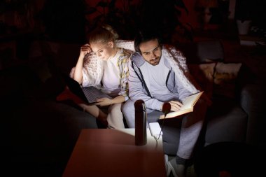 Akşam rutini. Genç çift kanepede oturuyor, dizüstü bilgisayarda çalışıyor ve evde oturma odasında kitap okuyor. Elektrik yok, elektrik kesintisi. Elektrik kesintisi ve aile toplantısı kavramı.