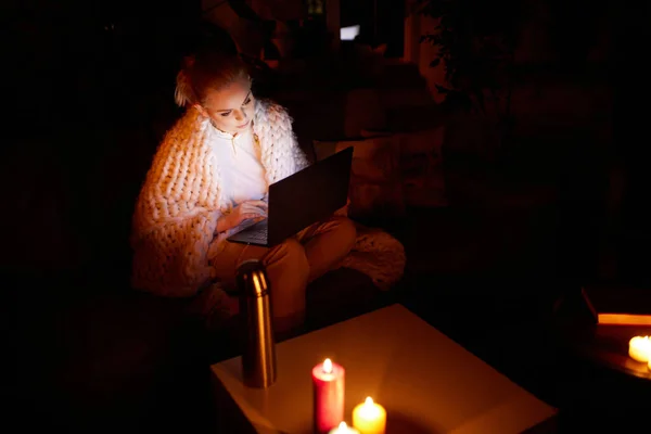 Cozy evening without electricity. Young beautiful girl sitting on sofa in warm plaid at home in the evening and working on laptop with candle light. Blackout. Concept of power outage