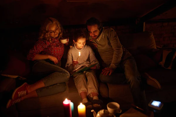 Cozy family evening. Mother, father and little girl sitting on sofa without electricity and reading book with candle light. Blackout. Hobby, leisure time. Concept of power outage, adjusting