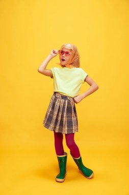 Vay canına. Pozitif bakış. Güzel küçük bir kız, şık giyinmiş bir çocuk duygusal olarak sarı stüdyo arka planında poz veriyor. Çocukluk kavramı, duygular, eğlence, moda, yaşam tarzı, yüz ifadesi