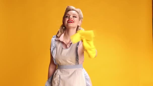 笑个不停漂亮的时尚女孩 穿着复古围裙的家庭主妇 带着橡胶手套在黄色的工作室背景上做清洁 复古时尚的概念 吸引力 平底锅风格 — 图库视频影像