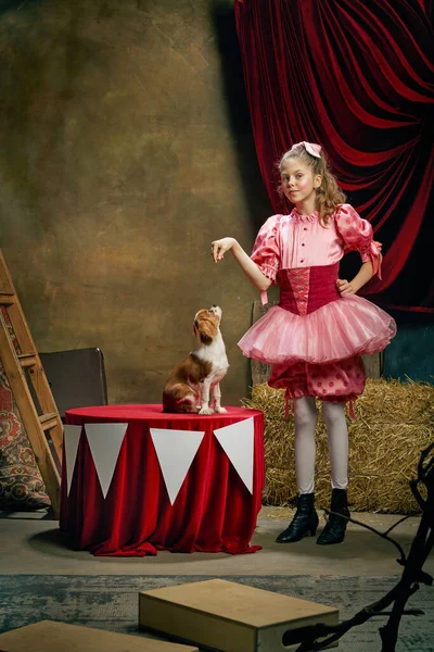 Making performance with dog. Beautiful little girl in festive pink dress posing over dark vintage circus background. Concept of retro circus, holidays, dreams, art, fashion, vintage style