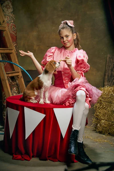 Feeding dog. Beautiful little girl in festive pink dress posing over dark vintage circus background. Concept of retro circus, holidays, dreams, art, fashion, vintage style
