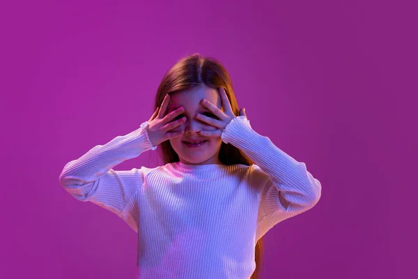 Hide and seek game. Portrait of little girl, child in casual clothes posing covering eyes with hands over purple studio background in neon light. Concept of childhood, emotions, fashion, lifestyle