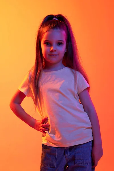 Calm smart kid. Portrait of little girl, child in casual clothes posing over gradient orange background in neon light. Concept of childhood, emotions, fashion, lifestyle, facial expression
