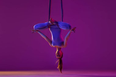 Upside down. Young flexible girl doing aerial yoga, training, swinging and meditating over purple studio background in neon light. Concept of fitness, sportive lifestyle, healthcare, strength