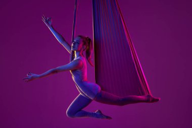 Positive vibes and relaxation. Young flexible girl doing aerial yoga, training over purple studio background in neon light. Concept of fitness, health, strength, aerial yoga, anti-gravity yoga