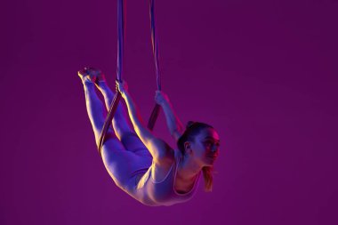Relaxing activity. Young flexible girl doing aerial yoga, training over purple studio background in neon light. Concept of fitness, sportive lifestyle, health, strength, aerial yoga, anti-gravity yoga