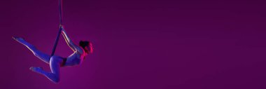 Stretching. Young flexible girl doing aerial yoga, training over purple studio background in neon light. Concept of fitness, health, aerial yoga, anti-gravity yoga. Banner, flyer. Copy space for ad