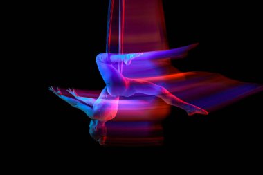 Young flexible girl doing aerial fly yoga, training over black studio background in neon with mixed lights. Concept of fitness, sportive lifestyle, health, strength, aerial yoga, anti-gravity yoga