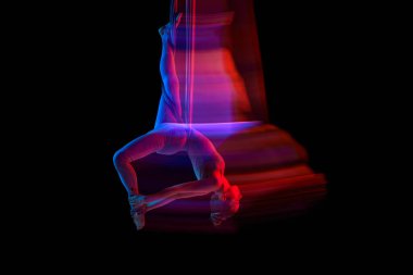 Stretching. Young flexible girl doing aerial yoga, training over black studio background in neon with mixed lights. Concept of fitness, sportive lifestyle, health, aerial yoga, anti-gravity yoga