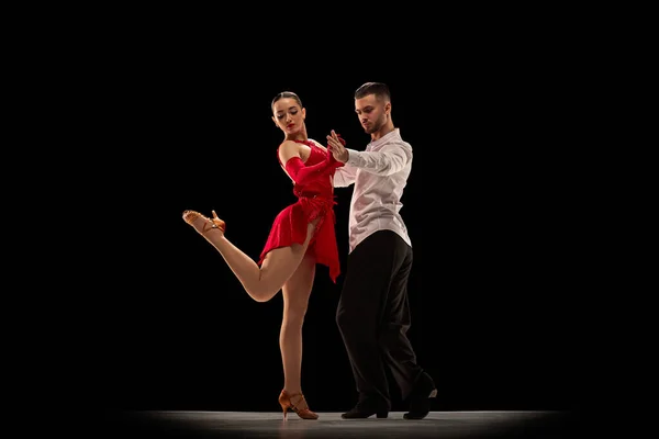 Professional dancers. Performance. Young man and woman dancing classical tango, ballroom over black background. Concept of hobby, lifestyle, action, beauty of movements, emotions, fashion, art