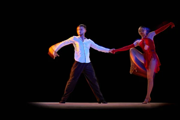 Man and woman, professional dancers in stylish stage costumes performing ballroom, tango over black background with mixed neon lights. Concept of lifestyle, action, beauty of movements, emotions, art