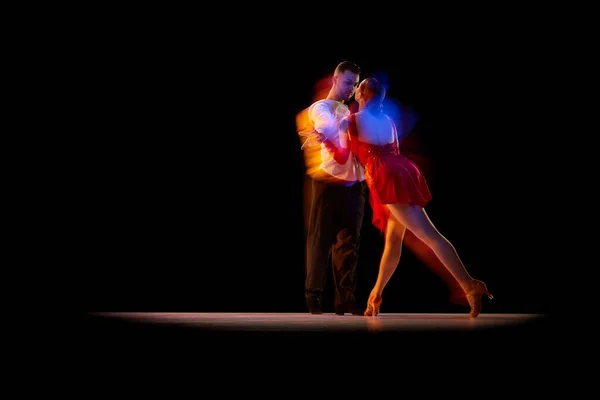 Man and woman, professional dancers in stylish stage costumes performing ballroom, tango over black background with mixed neon lights. Concept of hobby, lifestyle, action, beauty of movements, art