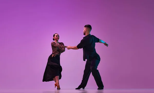 Man and woman, professional dancers in stylish stage costumes performing tango over purple background on neon lights. Concept of hobby, lifestyle, action, beauty of movements, emotions, fashion, art