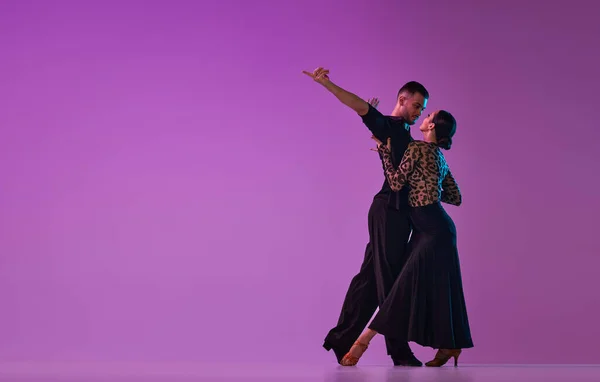 Love. Man and woman, professional dancers in stylish stage costumes performing tango over purple background on neon lights. Concept of lifestyle, action, beauty of movements, emotions, fashion, art