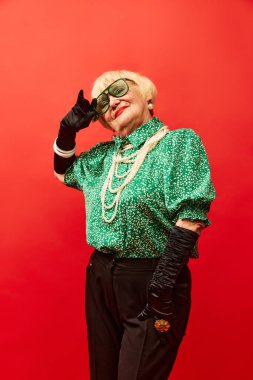Extraordinary lady. Beautiful old woman, grandmother in stylish clothes posing over red studio background. Concept of age, fashion, lifestyle, emotions, facial expression