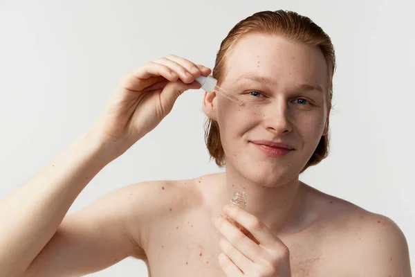 Young man taking care after skin. Redhead model posing against grey studio background. Applying face serum for hydration. Concept of mens health, body and skin care, hygiene and male cosmetology