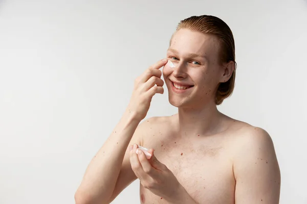 Young man taking care after skin. Redhead model posing against grey studio background. Applying moisturizing face cream. Concept of mens health, body and skin care, hygiene and male cosmetology