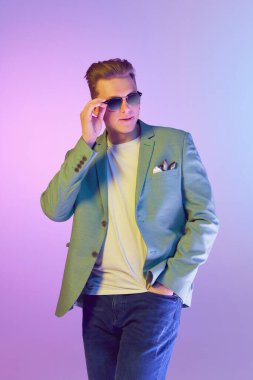 Charismatic male look. Young handsome man in stylish jacket and trendy sunglasses posing over purple background in neon light. Concept of emotions, lifestyle, youth, modern fashion. Ad
