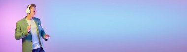 Young handsome man in stylish jacket posing, listening to music and dancing over purple background in neon light. Concept of emotions, lifestyle, youth, modern fashion. Banner. Copy space for ad