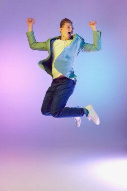 Feeling happiness. Energy burst. Young handsome man in stylish jacket posing, jumping over purple background in neon light. Concept of emotions, lifestyle, youth, modern fashion. Ad